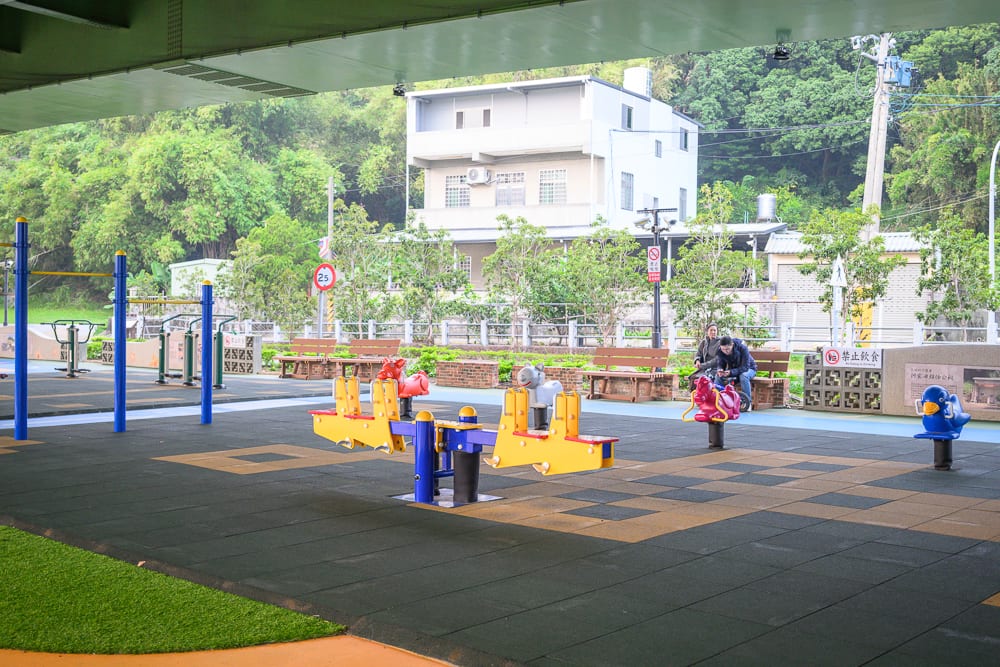苗栗滑步車公園｜高架橋下、遊樂場、彩色地墊、翹翹板、搖搖馬、木椅、綠樹、白色民宅、行人。