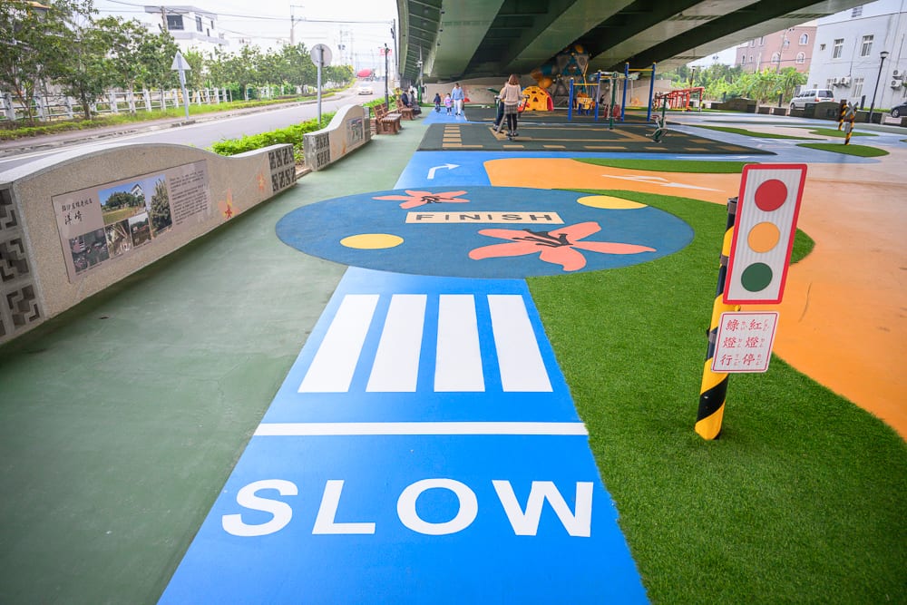 苗栗滑步車公園｜高架橋下、道路、斑馬線、紅綠燈、遊樂場、行人、人行道、草皮，藍色與黃色塗裝地面印有白色SLOW字樣與花朵圖案，遠處可見住宅建築與路樹。