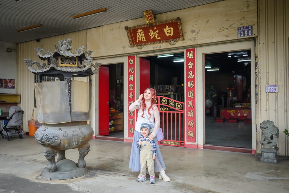 桃園白蛇廟｜寺廟，紅柱，香爐，招牌，女性，孩童，鐵皮屋。紅字匾額寫著白蛇廟，前方立著大型金屬香爐。紅衣女子與戴帽男童站在門口，兩側貼著對聯，門框內可見神桌與燈管照明。