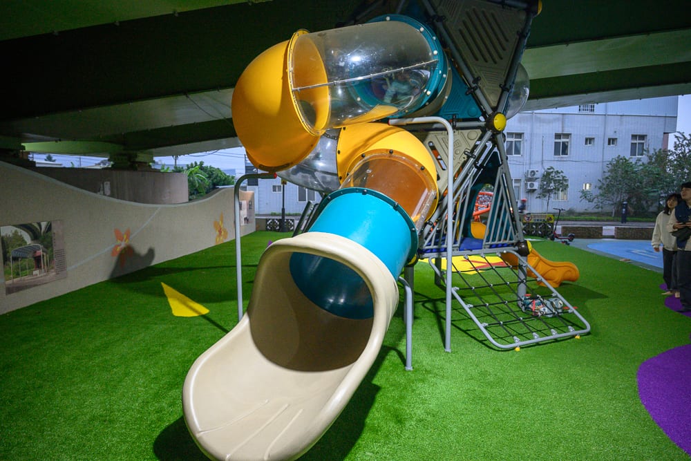 苗栗滑步車公園｜高架橋下的人工草皮兒童遊樂場，大型塑膠溜滑梯、透明管狀滑道、攀爬網、牆面裝飾，右側站立兩名行人，人工照明均勻打亮。