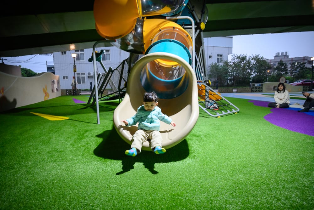 苗栗滑步車公園｜高架橋下公園，遊具，滑梯，兒童，人工草皮，人行道，建築物。幼童從黃色塑膠管狀滑梯滑下，穿著淺色外套，前方為綠色人工草皮鋪面，背景可見高架橋結構與遠處建築，右側地面坐有成人。