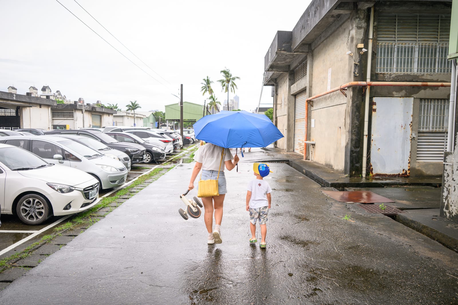 高雄親子遊樂園區完整指南|室內遊具與軌道體驗玩法 8 高雄親子遊樂園區|街道、汽車、倉庫、撐傘成人、小孩,濕漉漉的柏油路面映出微弱反光,兩旁停放多輛白色與黑色車輛,高大廠房牆面斑駁,陰天光線均勻。