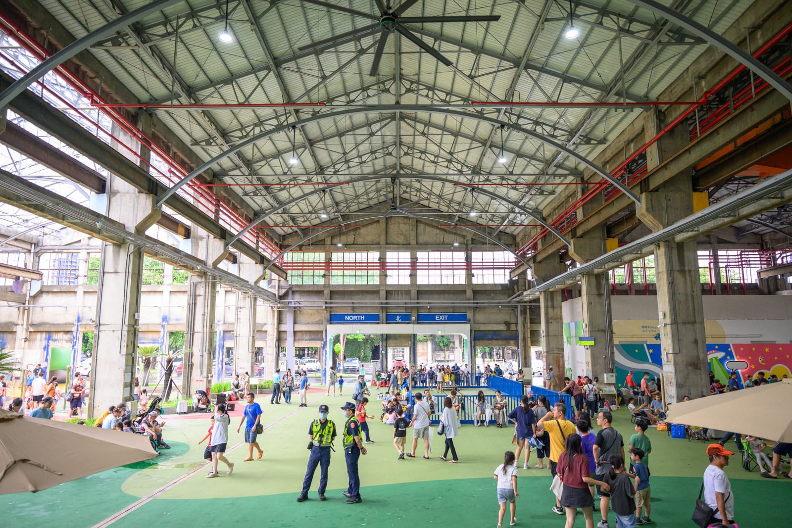 高雄親子遊樂園區完整指南|室內遊具與軌道體驗玩法 6 高雄親子遊樂園區|挑高鋼構廠房內部空間,大片綠色地面鋪面,水泥柱列支撐拱形屋頂。人群分散於室內活動,兩名穿制服員警站在中央,遠處設有出口標示與窗戶透光。