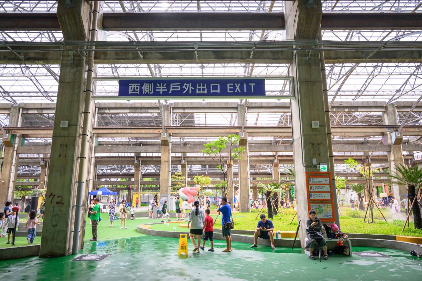 高雄親子遊樂園區完整指南|室內遊具與軌道體驗玩法 5 高雄親子遊樂園區|混凝土柱、網格屋頂、藍底白字標示牌、綠地、行人、座椅,挑高的工業風半戶外空間,柱列支撐著透光屋頂,地面覆蓋人工草皮與綠色鋪面,許多遊客站立、行走或坐在柱邊休憩。