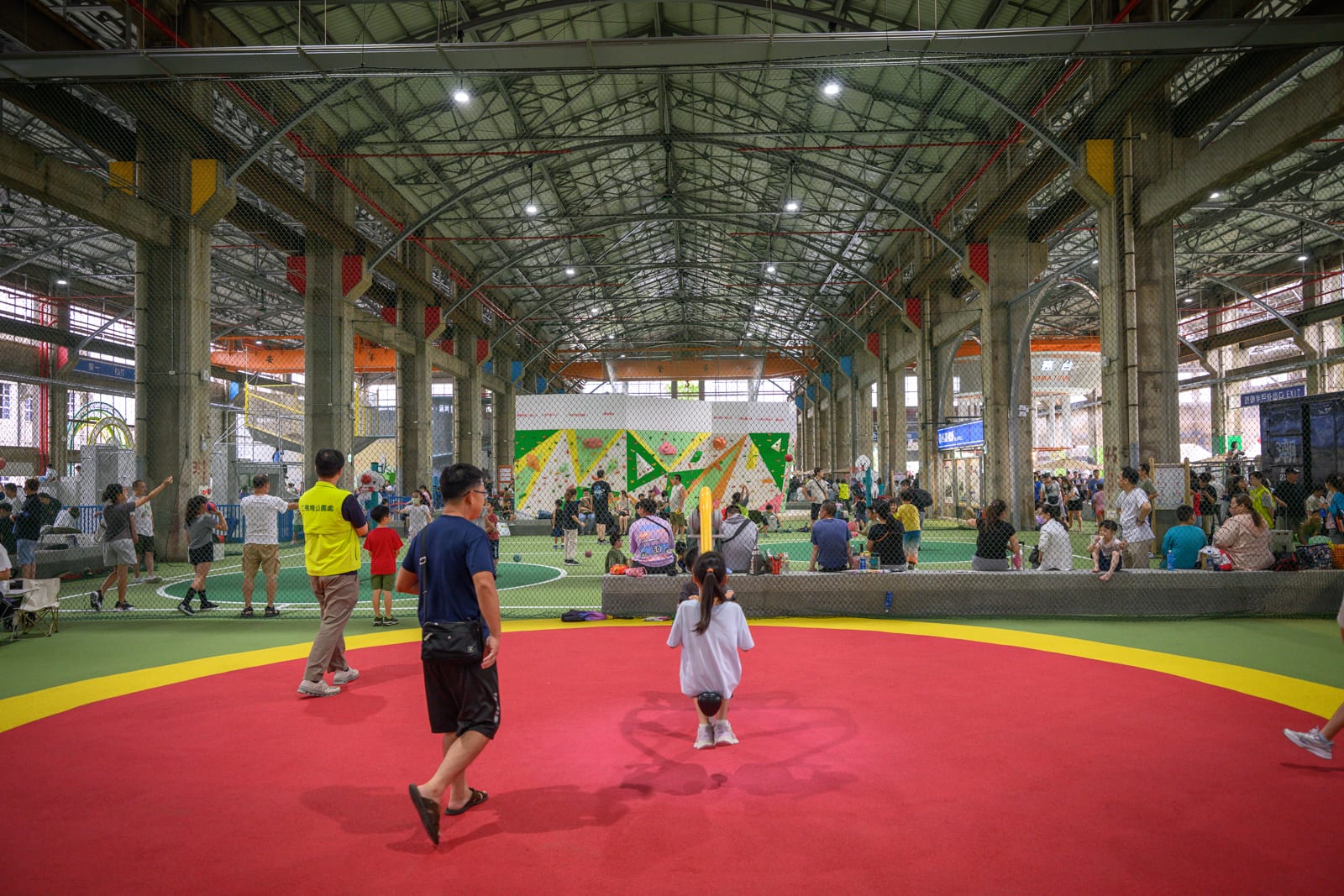 高雄親子遊樂園區完整指南|室內遊具與軌道體驗玩法 11 高雄親子遊樂園區|挑高鋼架室內,大型攀岩牆,紅黃綠相間運動地墊,防護網,人群,混凝土柱,球場。屋頂吊燈照明,人們在場地內走動與休憩。
