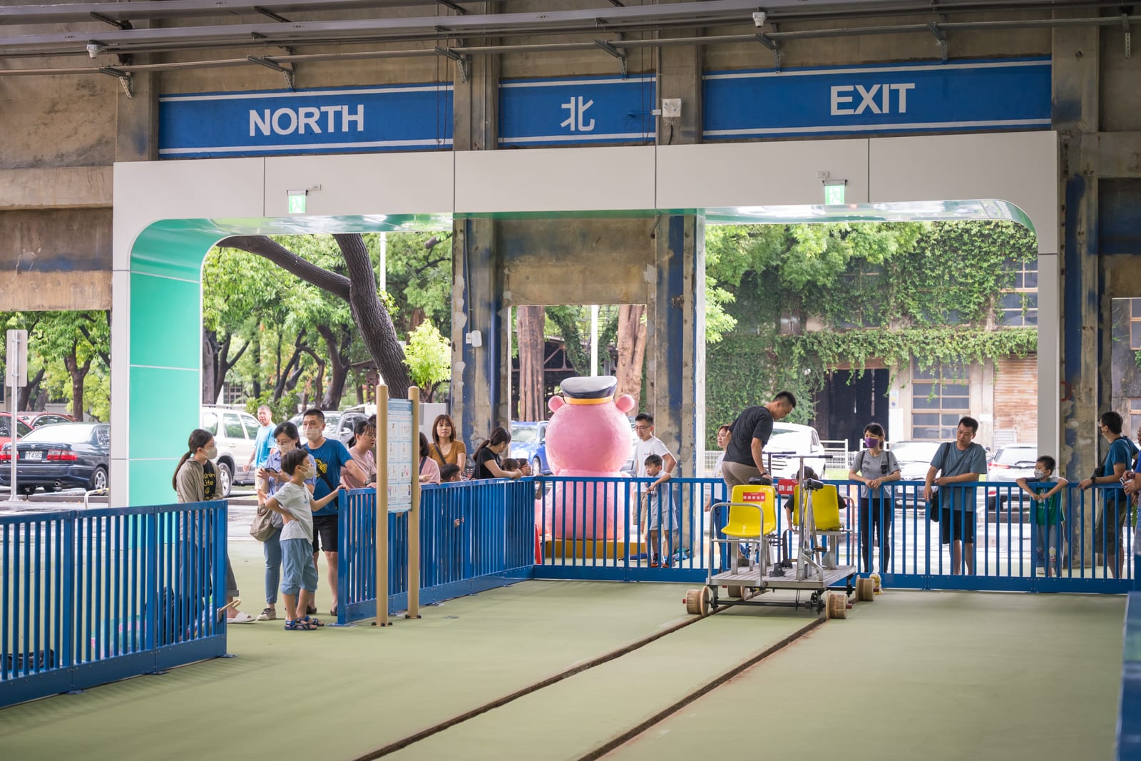 高雄親子遊樂園區完整指南|室內遊具與軌道體驗玩法 4 高雄親子遊樂園區|車站、軌道、欄杆、人群、招牌,室內候車月台邊設有藍色金屬柵欄,軌道上有小型手動台車,遊客聚集在柵欄兩側觀看,上方懸掛藍底白字的英文與中文出口標示。