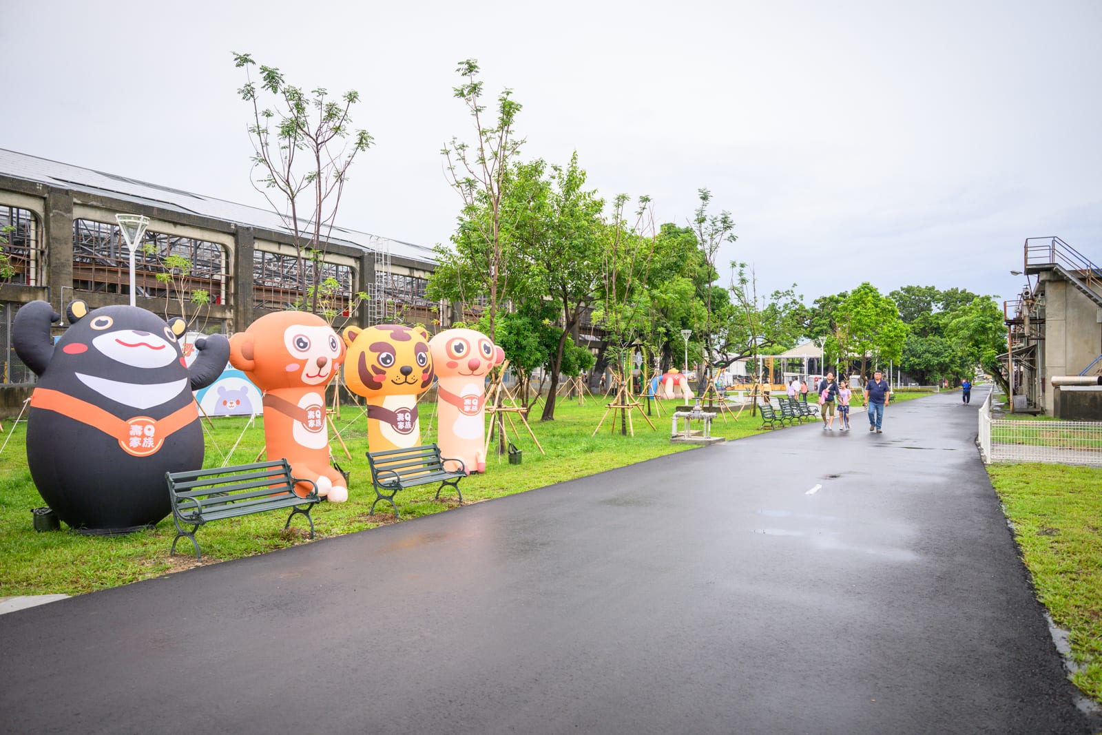 高雄親子遊樂園區完整指南|室內遊具與軌道體驗玩法 7 高雄親子遊樂園區|柏油路面、氣球、長椅、草皮、樹木、倉庫。戶外園區草地上並排立著四個造型充氣玩偶,左側有兩張黑色鐵架長椅,右側寬敞道路上有少數行人漫步,背景為低矮廠房建築與林木。