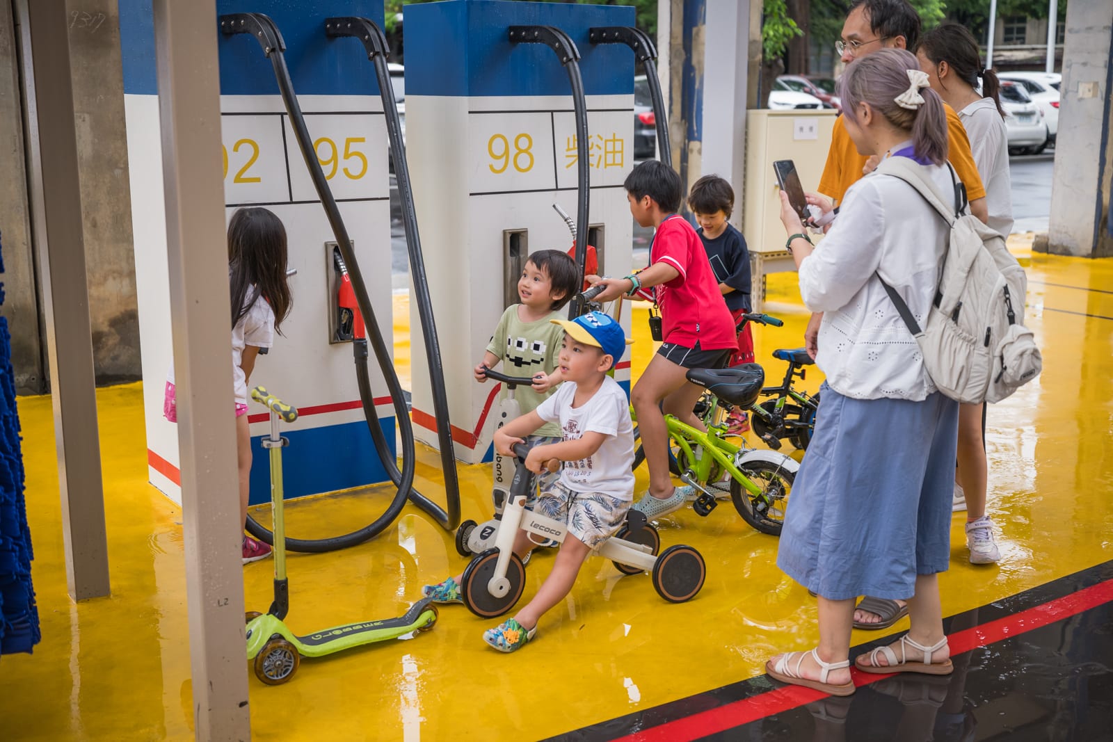 高雄親子遊樂園區完整指南|室內遊具與軌道體驗玩法 2 高雄親子遊樂園區|加油站,兒童,滑步車,腳踏車,大人,手機。孩子們在模仿加油操作,大人在一旁拍照記錄,濕漉漉的黃色地坪反射著光線,四周停放著車輛。
