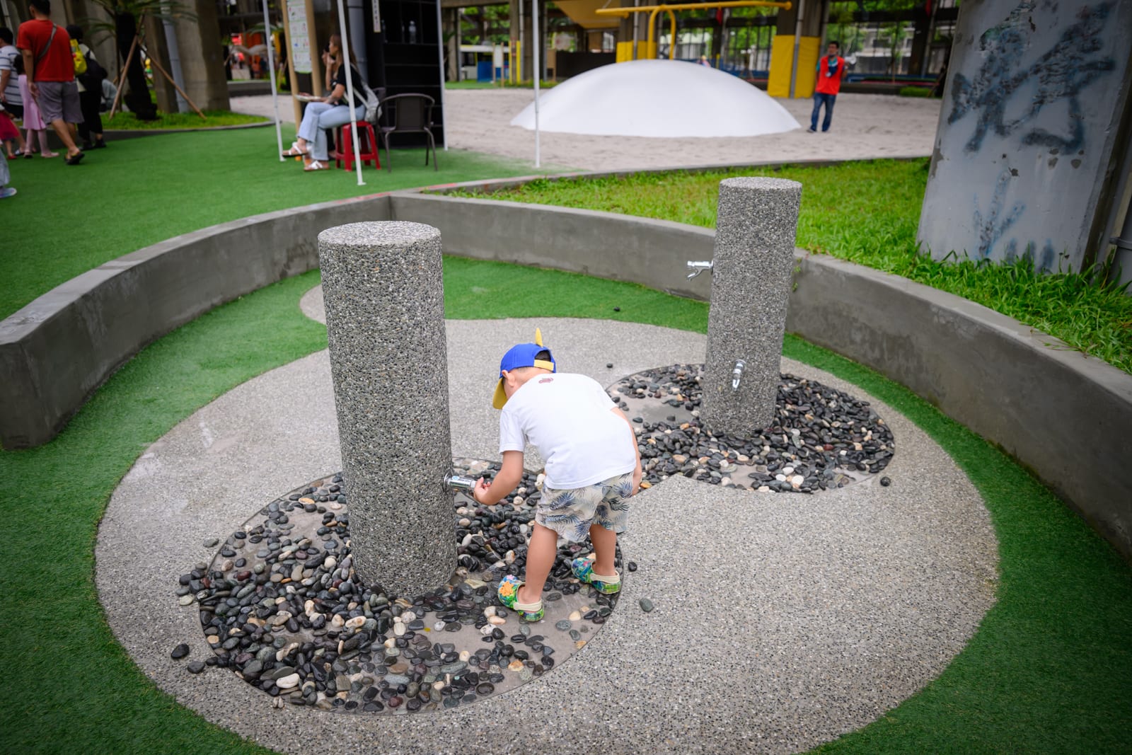 高雄親子遊樂園區完整指南|室內遊具與軌道體驗玩法 10 高雄親子遊樂園區|兒童遊樂場、圓形水池、石柱、鵝卵石、人工草皮。戴帽男童正彎腰操作石柱上的水龍頭,後方水泥花圃與白色半圓形設施,遠處有行人與桌椅。