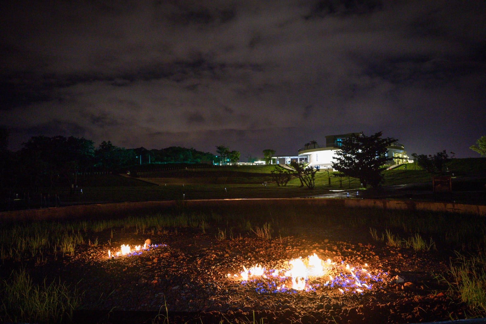 屏東恆春出火地質公園完整指南|天然氣火焰與夜間賞火玩法 3 屏東恆春出火地質公園|夜間草地,火源,建築,遠山,陰暗天空。前景凹陷處燃燒著帶有橘黃與藍色火焰的泥地,中景為大片黑暗草皮與疏落樹木,遠方山坡上有亮燈的現代建築,上方為佈滿雲層的夜空。