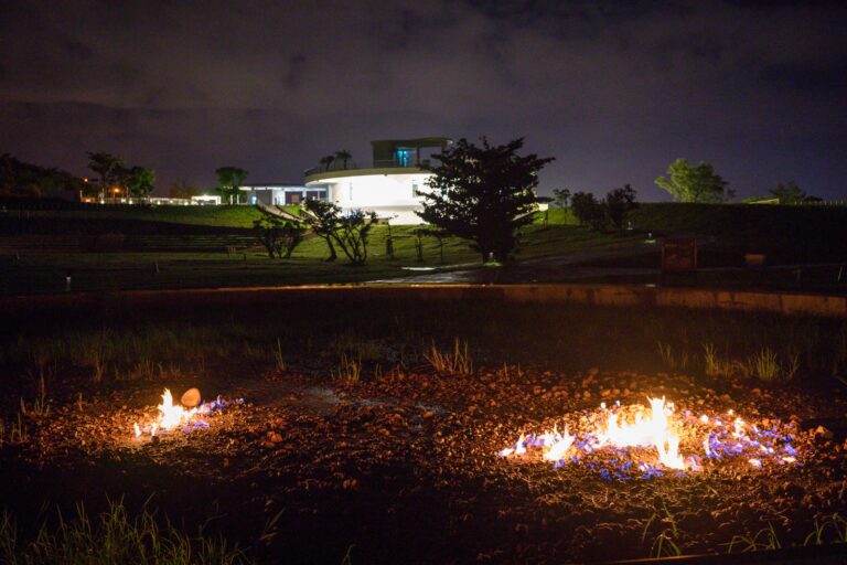 屏東恆春出火地質公園完整指南|天然氣火焰與夜間賞火玩法 15 屏東恆春出火地質公園|草地、建築、火焰、夜空。夜間戶外草地上,兩處地面正燃燒著橘藍色火焰,火光映照周圍土壤與稀疏雜草。遠方中景為一座白色圓形現代建築,散發明亮燈光。背景為暗灰色夜空與遠山剪影。