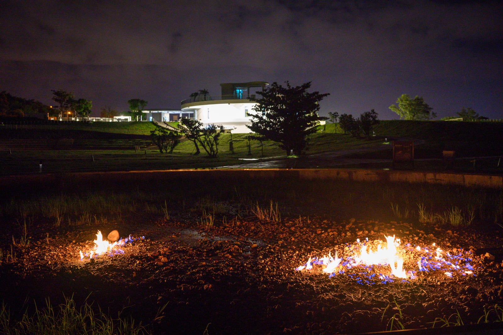 屏東恆春出火地質公園完整指南|天然氣火焰與夜間賞火玩法 1 屏東恆春出火地質公園|草地、建築、火焰、夜空。夜間戶外草地上,兩處地面正燃燒著橘藍色火焰,火光映照周圍土壤與稀疏雜草。遠方中景為一座白色圓形現代建築,散發明亮燈光。背景為暗灰色夜空與遠山剪影。