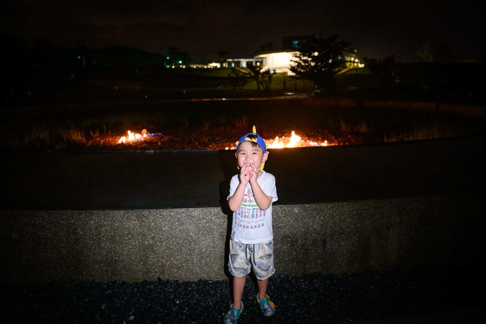 屏東恆春出火地質公園完整指南|天然氣火焰與夜間賞火玩法 7 屏東恆春出火地質公園|男孩、帽子、短褲、石牆、夜景、火焰、草地、建築,黑夜戶外場景。男孩站在矮石牆前,頭戴藍黃色鴨舌帽與白色上衣,雙手捧著臉頰微笑。背景遠處暗色草地上有兩處跳動橘色火焰,更遠方有亮燈的低矮現代建築與樹木剪影。