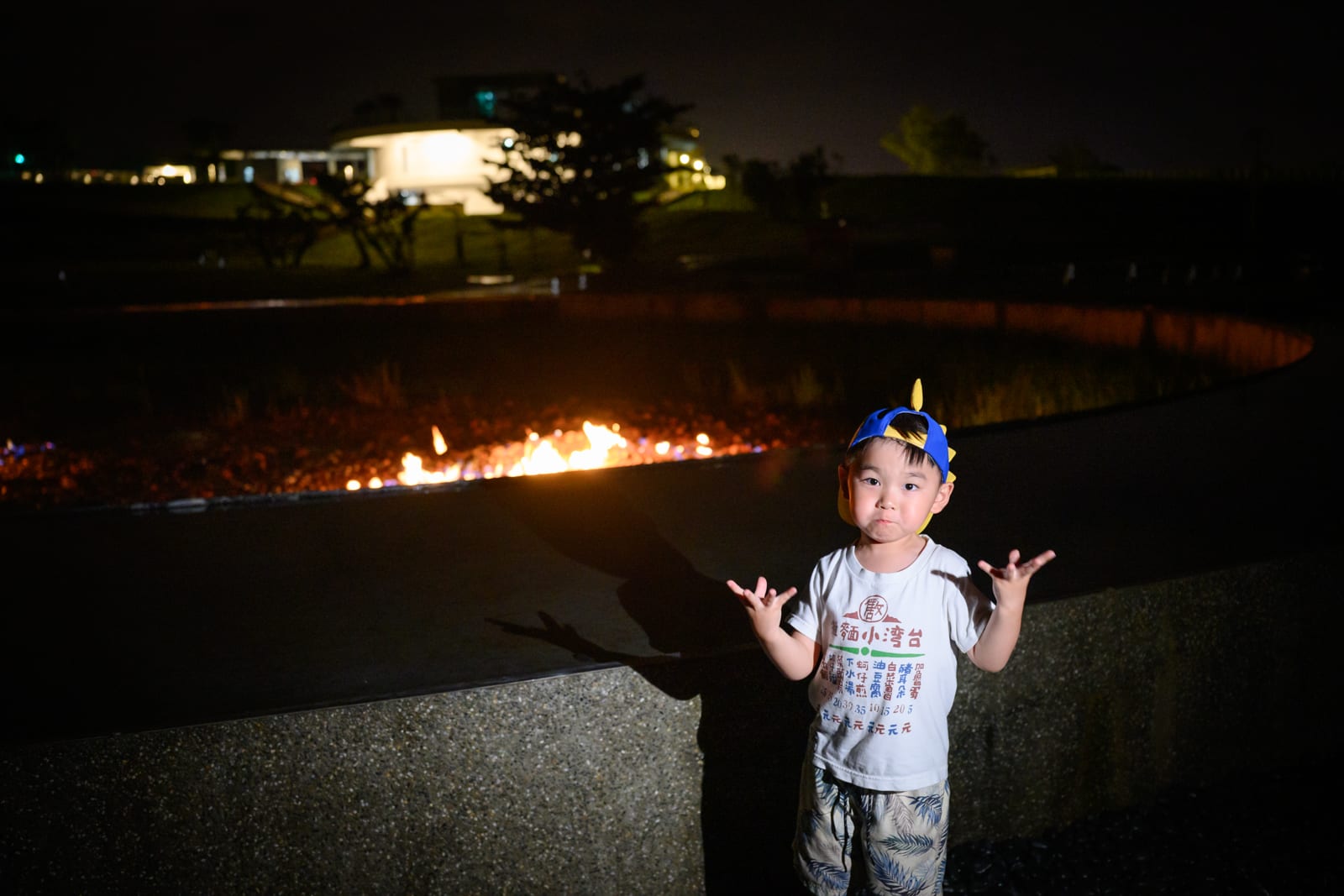屏東恆春出火地質公園完整指南|天然氣火焰與夜間賞火玩法 2 屏東恆春出火地質公園|夜間戶外,男童,帽子,短袖上衣,短褲,石牆,火焰,遠方燈光建築,草地。男童站在半圓形矮牆前,雙手張開比出動作,背後石槽內燃燒著橘黃色火焰,背景為黑夜與發光的建築物。