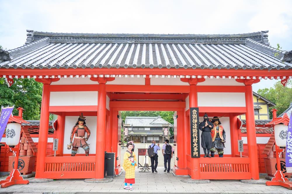 日本登別伊達時代村完整指南|江戶街景與庭園展示 8 日本登別-依達時代村|城門、寺廟、武士雕像、穿著和服的人、行人,灰色瓦片屋頂搭配紅柱白牆結構,兩側設有燈籠,中央通道可見遠方傳統建築,明亮天光下呈現日式古建築景觀。