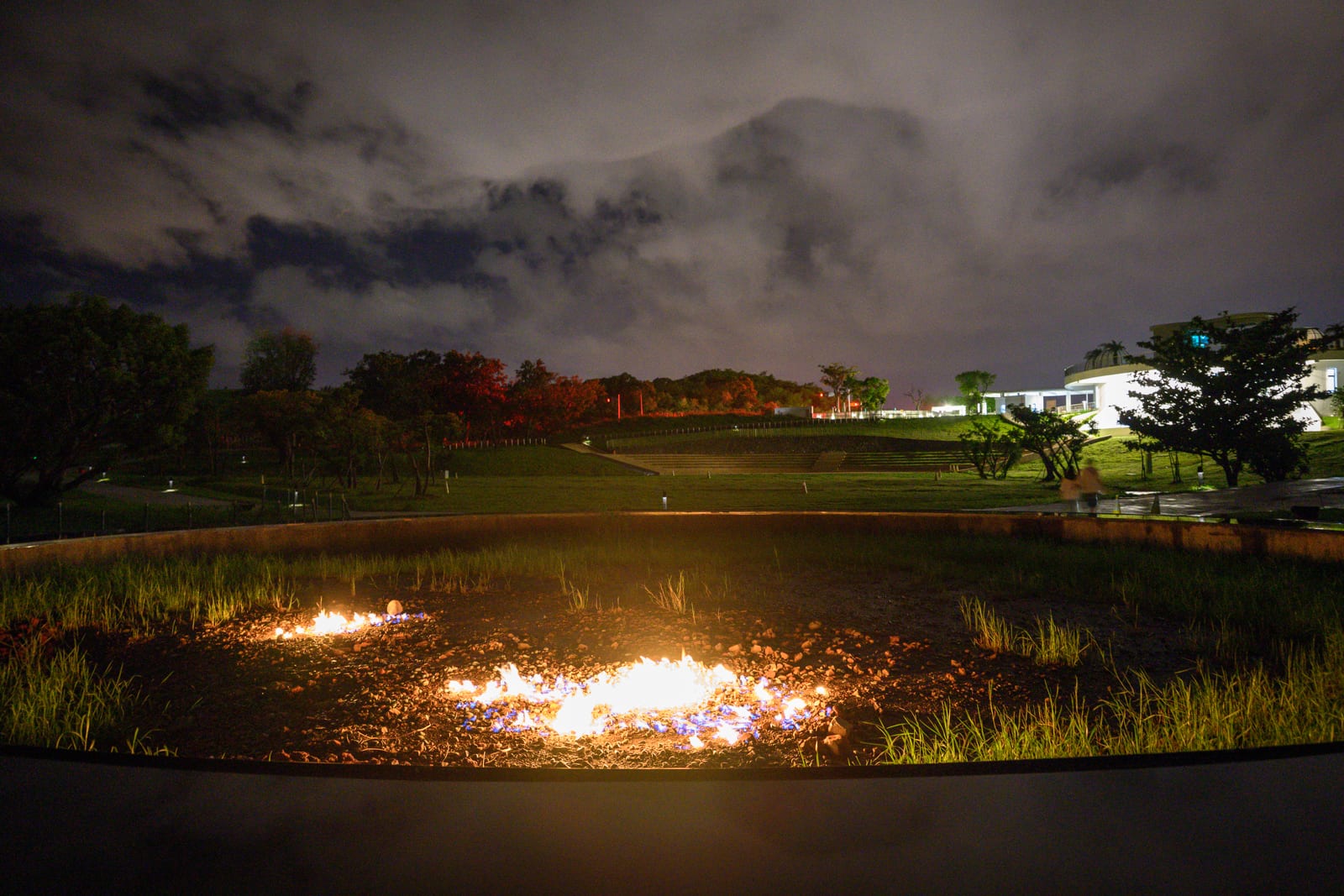 屏東恆春出火地質公園完整指南|天然氣火焰與夜間賞火玩法 8 屏東恆春出火地質公園|夜間草地、火焰、水池、樹木、低矮建築、雲層天空。凹陷窪地中燃燒著橘黃色火焰,照亮周邊綠草與土壤,遠方為起伏的暗色山丘與低矮現代建築,上空覆蓋著帶有縫隙的夜間雲層,微弱光線映照場景。
