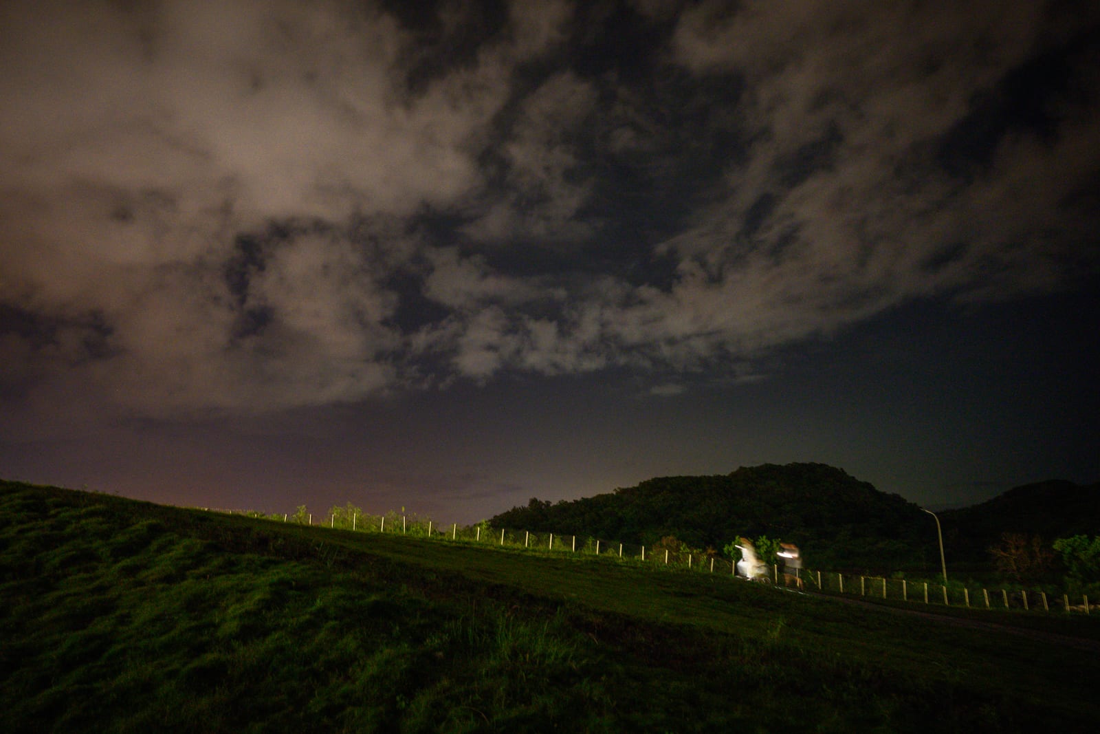 屏東恆春出火地質公園完整指南|天然氣火焰與夜間賞火玩法 6 屏東恆春出火地質公園|夜空、雲層、山丘、草坡、步道、護欄、路燈,夜間戶外草地步道上有人員移動殘影,遠方山頭輪廓清晰。