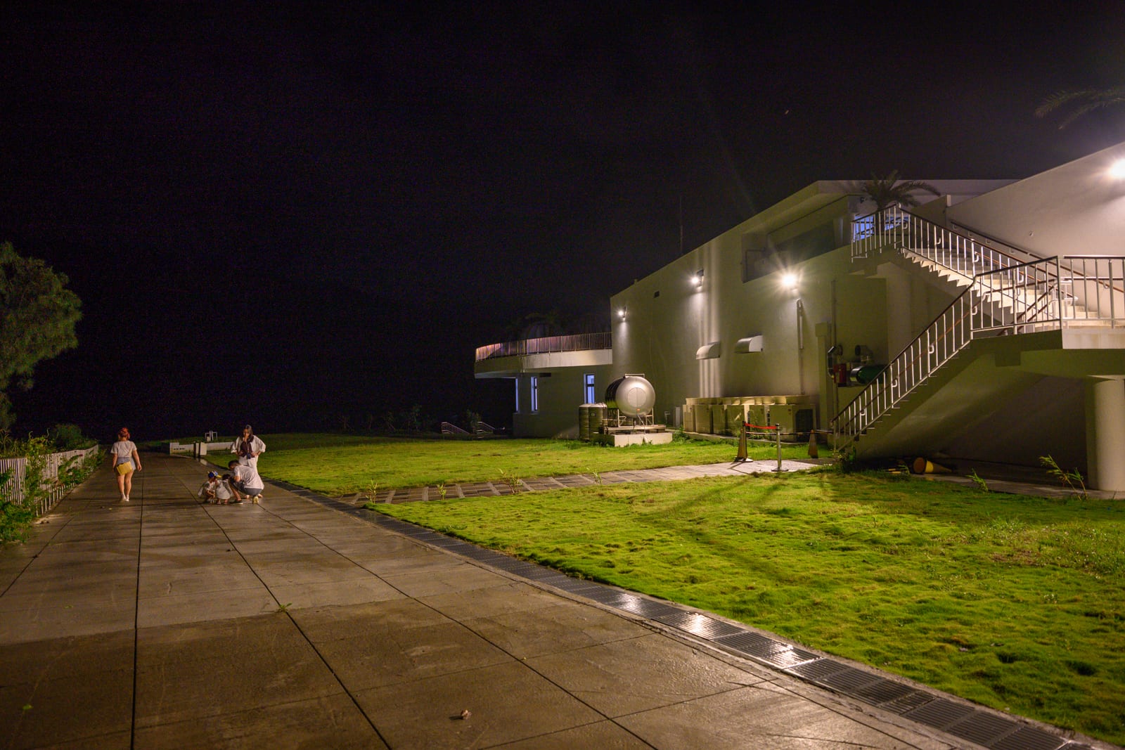 屏東恆春出火地質公園完整指南|天然氣火焰與夜間賞火玩法 12 屏東恆春出火地質公園|夜間建築、草地、行人、戶外階梯、步道,建築牆面嵌設多盞投射燈,照亮草皮與水泥地面,遠處黑夜與少數人物。