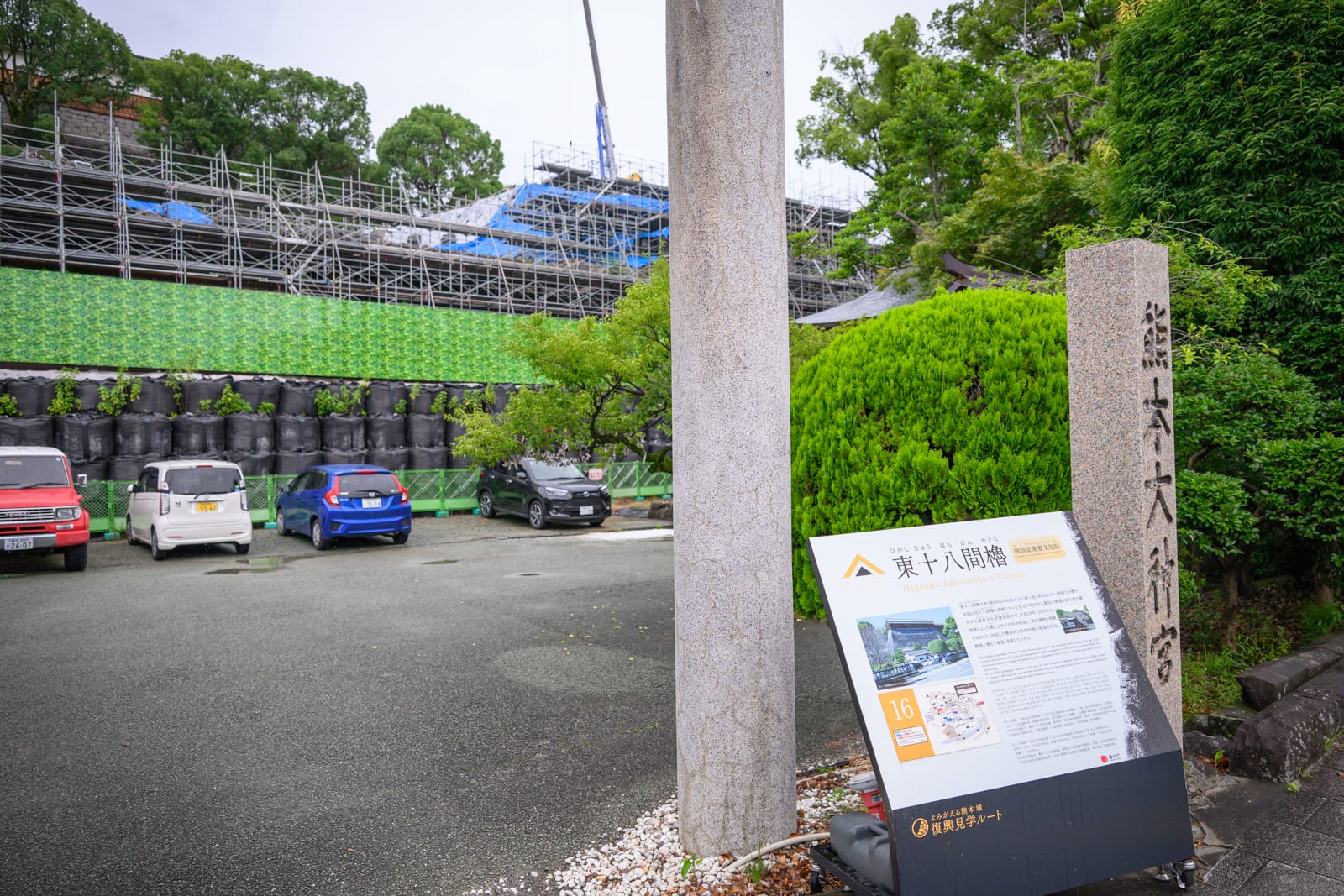 日本熊本-加藤神社｜石柱、告示牌、汽車、停車場、鷹架、綠樹。戶外停車場旁立有日文石碑與解說牌，背景為被施工鷹架與綠網覆蓋的建築物，多輛小型車停放於鋪面空地上。