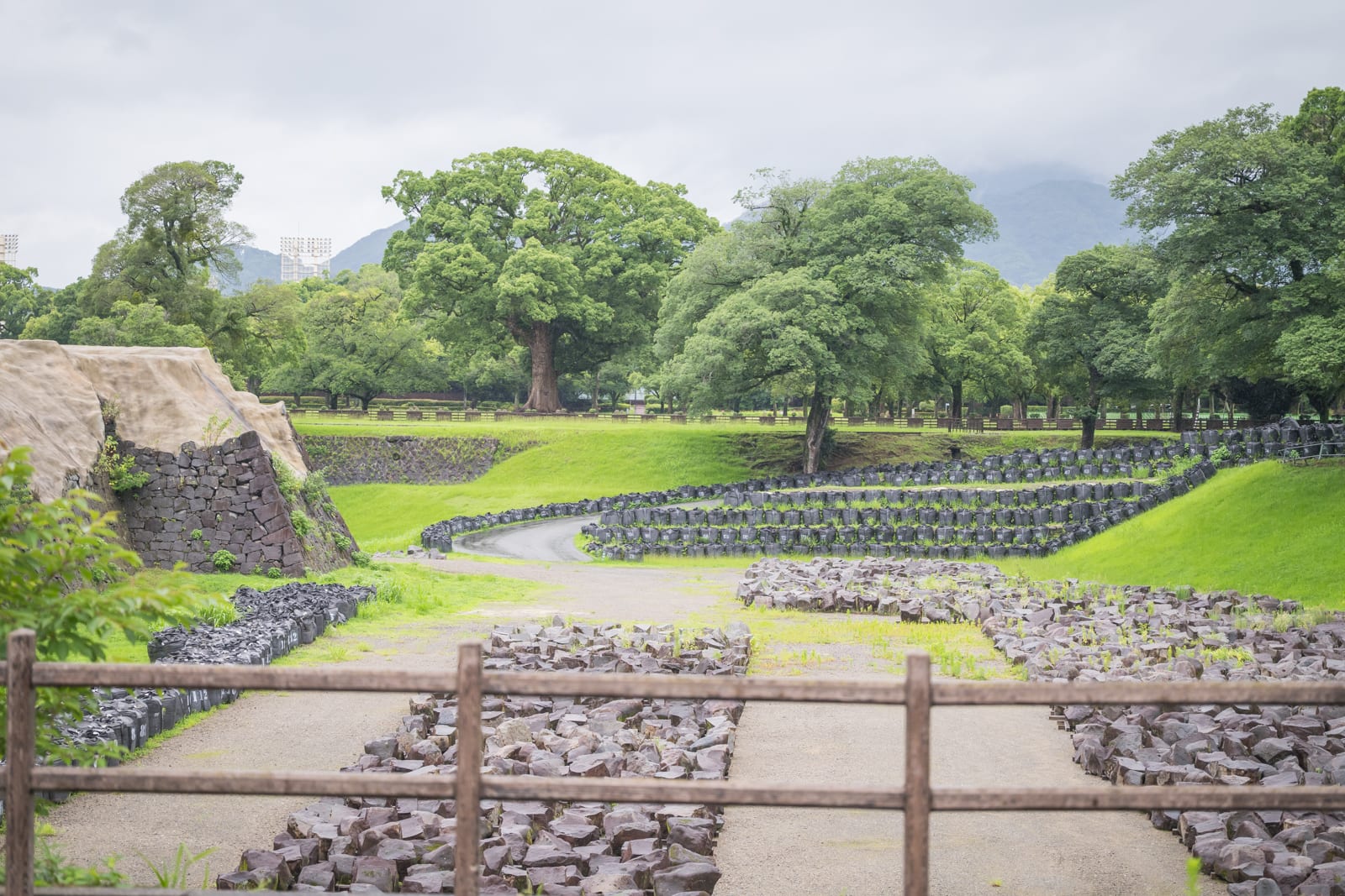 日本熊本-加藤神社｜木柵欄、石牆、草地、樹木、遠山、雲層，陰天光線下的公園戶外場景，遠處有巨大綠樹與平坦草坪，近景為排列整齊的石塊與泥土小徑。