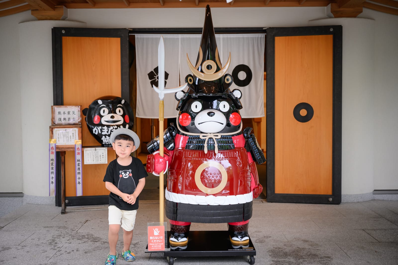 日本熊本-加藤神社｜建築物、木門、男孩、熊本熊立牌、武士盔甲、長槍。短髮男孩戴著帽子站立於巨型吉祥物雕像旁，雕像身穿紅色武士服裝並手持武器，背景為木造建築入口與布簾，地面為灰色磁磚。