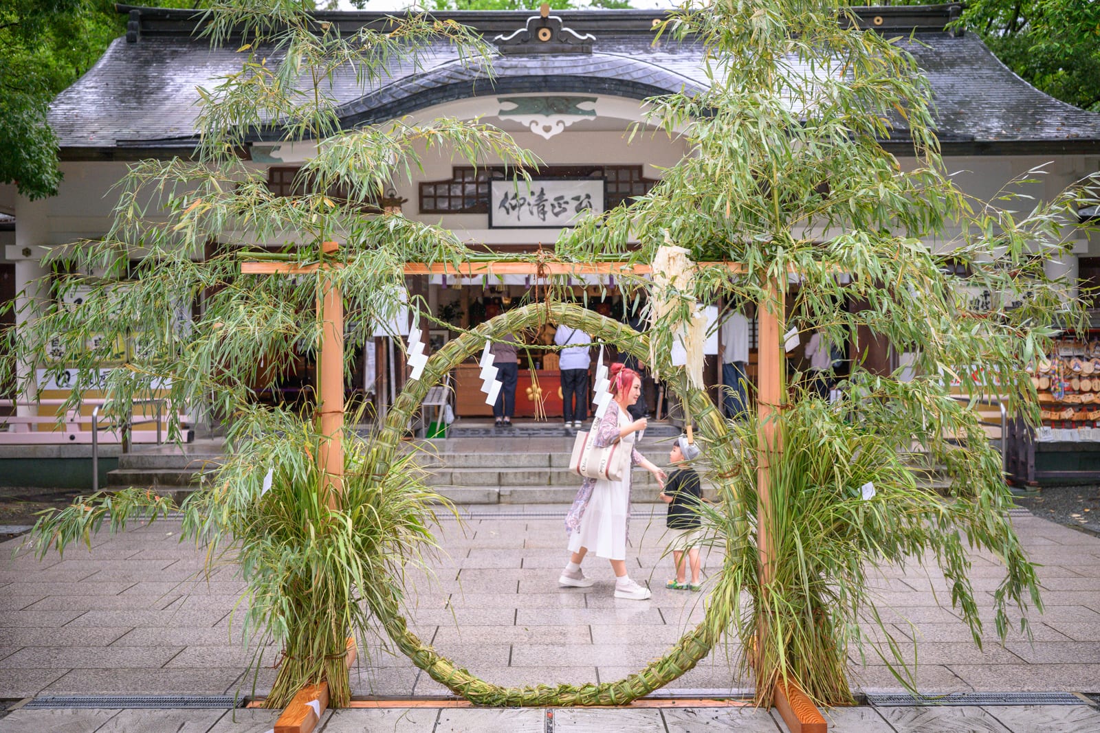 日本熊本-加藤神社｜寺廟、茅輪、竹子、女性、小孩，廣場石板地微濕，木造神社前設置草編圓形拱門，垂掛白色紙垂，綠竹枝葉茂密覆蓋框架，行人穿梭其間。