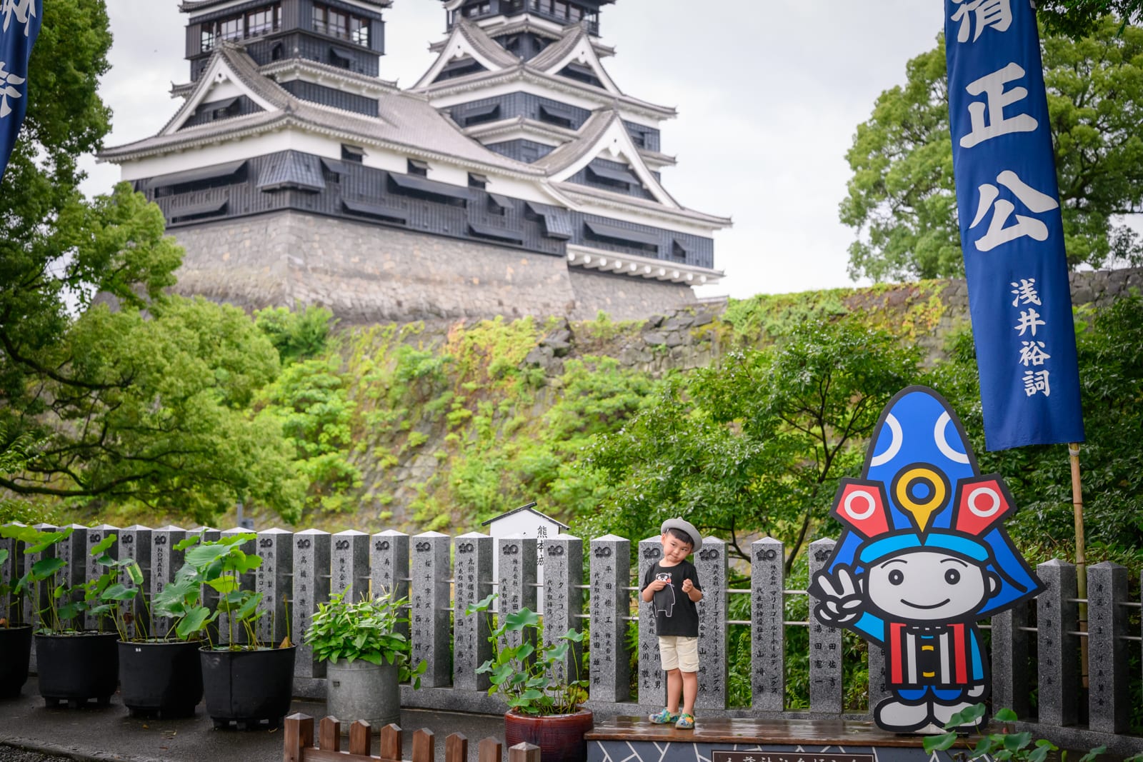 日本熊本-加藤神社｜城堡、護城牆、小男孩、吉祥物立牌、藍色旗幟、盆栽、石柱、綠樹。陰天光線下，遠處為層疊的黑瓦白牆城堡，下方石牆長滿綠意。石柱排列成矮牆，前方立著彩色吉祥物立牌與藍色長布旗，穿短袖短褲的小男孩站在石台上。