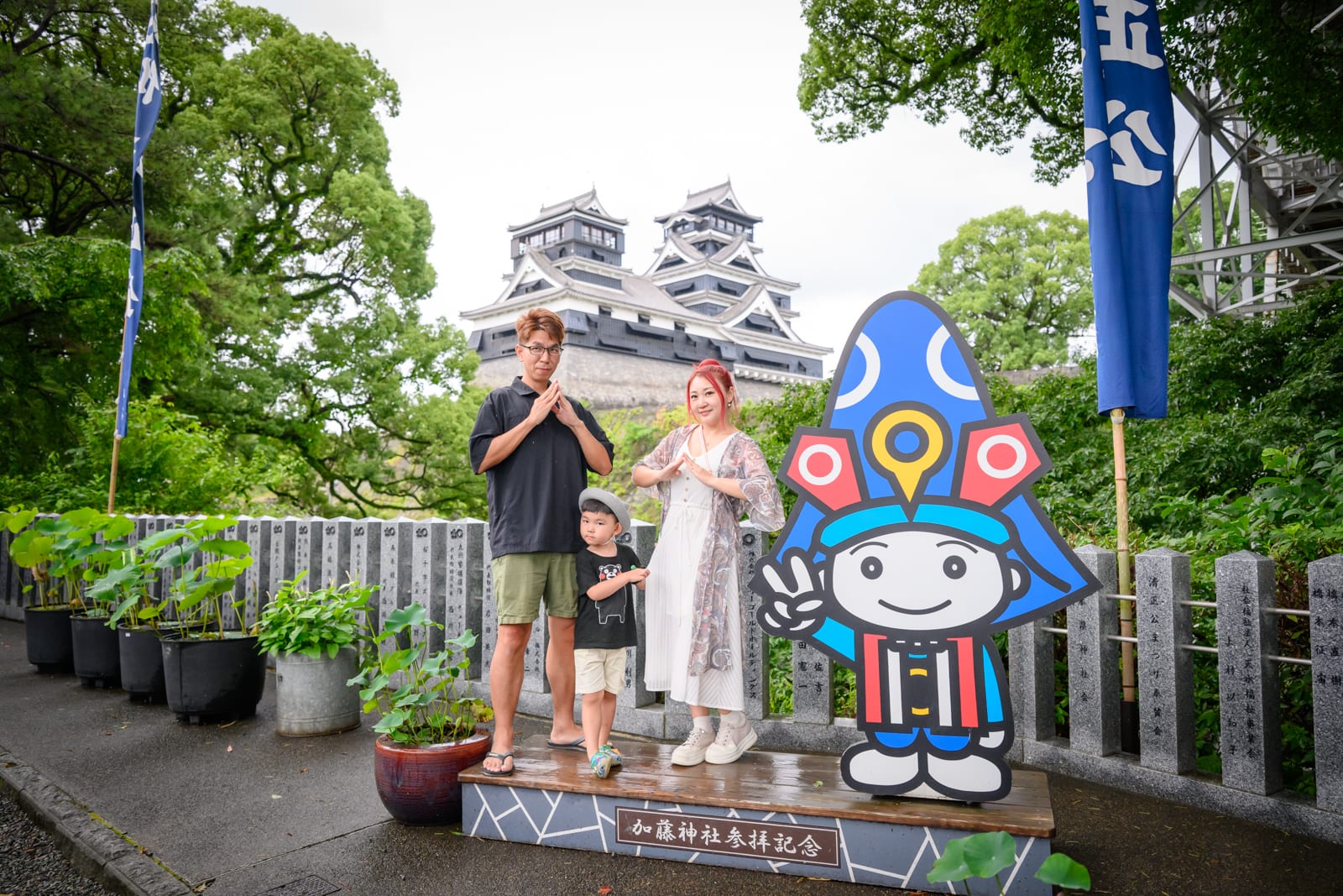 日本熊本-加藤神社｜一家三口與吉祥物立牌，城堡、石牆、旗幟，綠樹盆栽與石柱街道，陰天戶外廣場。
