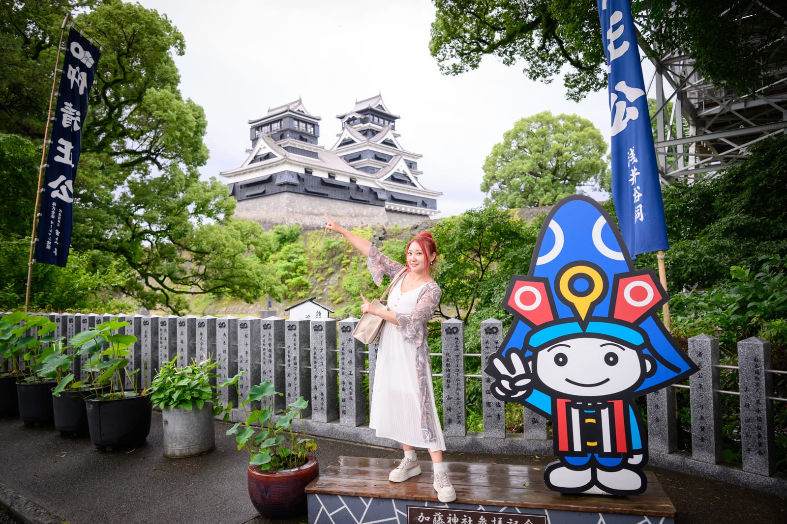 日本熊本-加藤神社｜城堡,旗幟,吉祥物,女子,護欄,植物。一名紅髮女子身穿白裙站在戶外，手持提袋並指向後方的黑色城堡。右側立著一個藍色人偶吉祥物，兩側插有日式布旗，前方有盆栽與石製護欄。