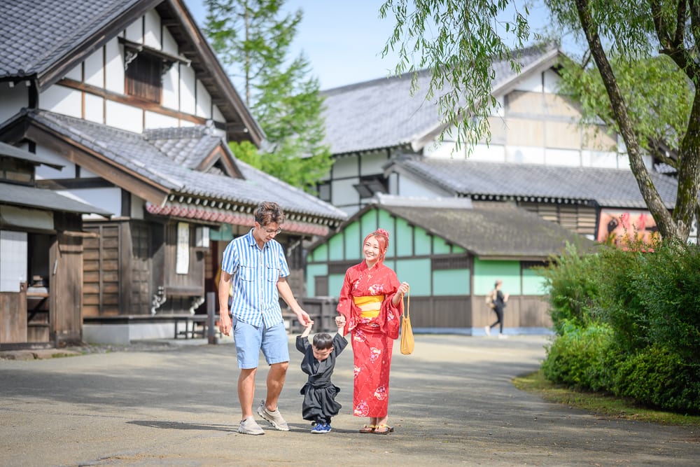 日本登別伊達時代村完整指南|江戶街景與庭園展示 11 日本登別-依達時代村|一家三口走在日式老街,父親穿條紋襯衫短褲,母親著紅色浴衣,牽著穿黑色服裝的小孩。背景為傳統黑瓦木造房屋、綠樹與泥土小路,頭頂枝葉垂拂。