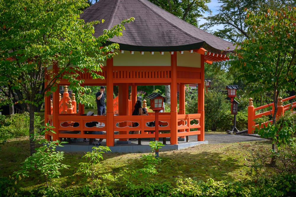 日本登別伊達時代村完整指南|江戶街景與庭園展示 12 日本登別-依達時代村|涼亭、寺廟、樹木、草皮,朱紅色六角涼亭座落綠意庭園中,屋頂深灰微尖,亭內有遊客站立與坐著交談,周圍環繞綠樹與草地,一側可見橘紅欄杆。