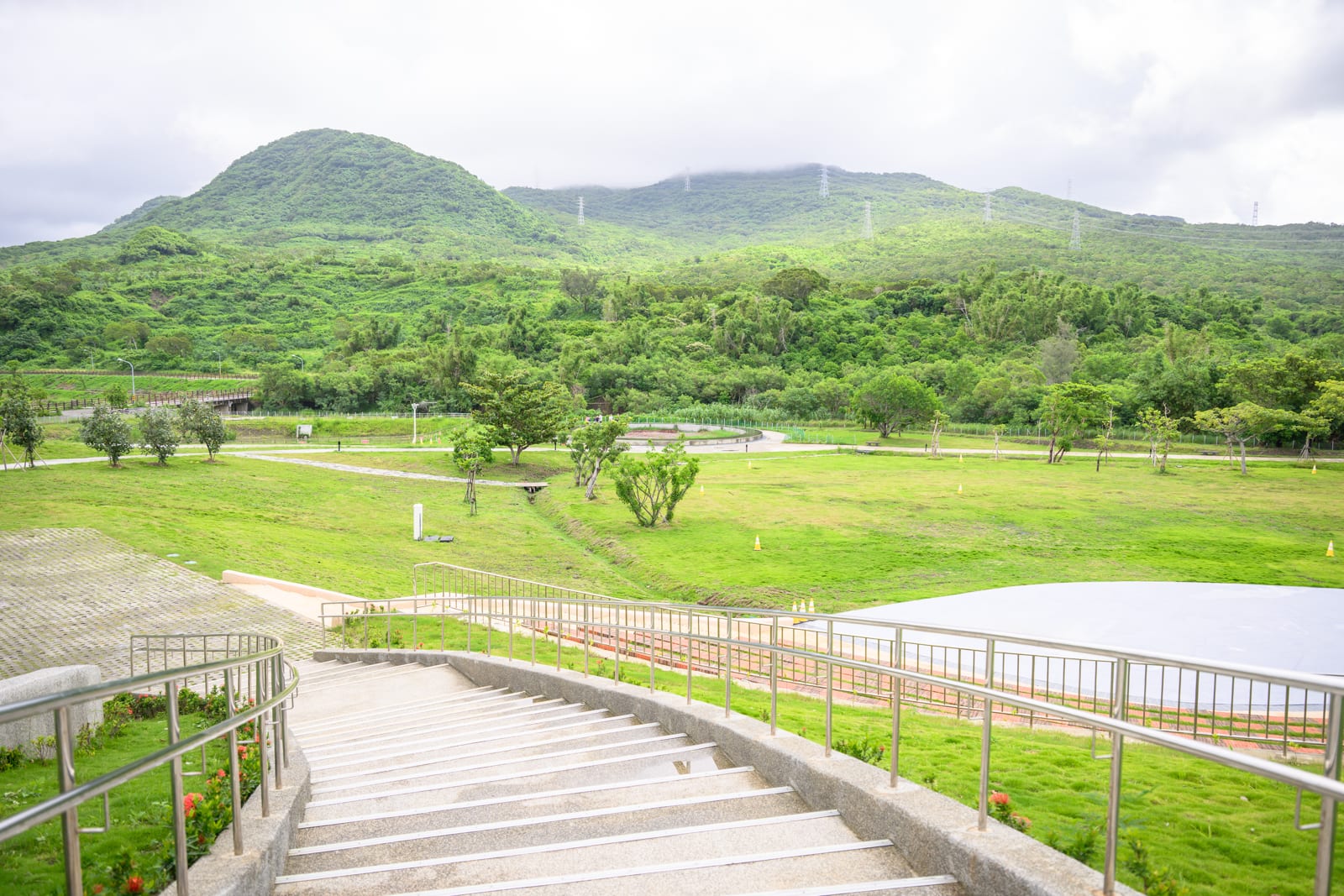屏東恆春出火地質公園完整指南|天然氣火焰與夜間賞火玩法 10 屏東恆春出火地質公園|階梯,草地,山丘,金屬扶手,雲霧,步道。向下延伸的石階設有金屬欄杆,前方為大片綠色草皮與稀疏樹木,遠方為層疊的綠意山巒,山頭壟罩薄霧,天空佈滿雲層。