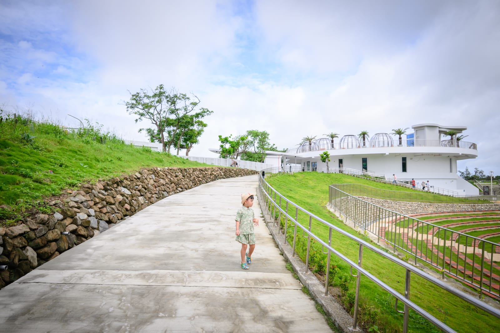 屏東恆春出火地質公園完整指南|天然氣火焰與夜間賞火玩法 5 屏東恆春出火地質公園|小孩、斜坡、石牆、草皮、白色建築、欄杆、天空,戶外階梯廣場旁的水泥步道上有戴帽幼童行走,遠方白色多層圓頂建築與綠地相鄰。