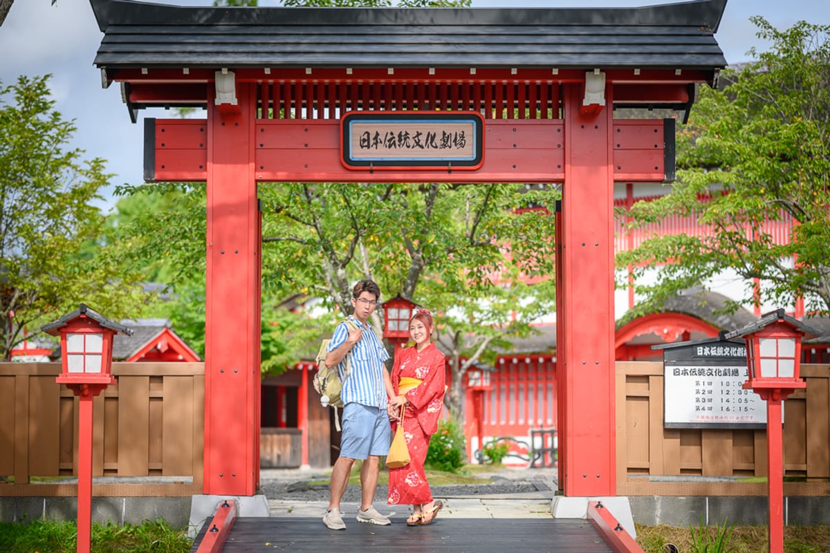日本登別伊達時代村完整指南|江戶街景與庭園展示 1 日本登別-依達時代村|木造鳥居、紅色燈籠、寺廟、橋、男女遊客、和服、綠樹,男女站立於紅色巨大傳統牌樓下方的石造小橋,牌樓上方懸掛黑框白字橫幅看板,兩側立有紅框白面直立燈柱,背景可見紅柱白牆建築群與綠意植被。