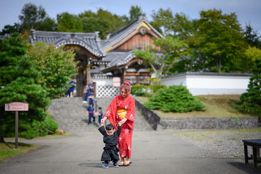 日本登別伊達時代村完整指南|江戶街景與庭園展示 6 日本登別-依達時代村|寺廟,紅衣女子,黑衣小孩,柏油路,石階,綠樹,木造建築。紅衣女子牽著黑衣小孩走在寺廟前的柏油路面上,背景為日式傳統建築與階梯,周圍環繞綠樹與草地。