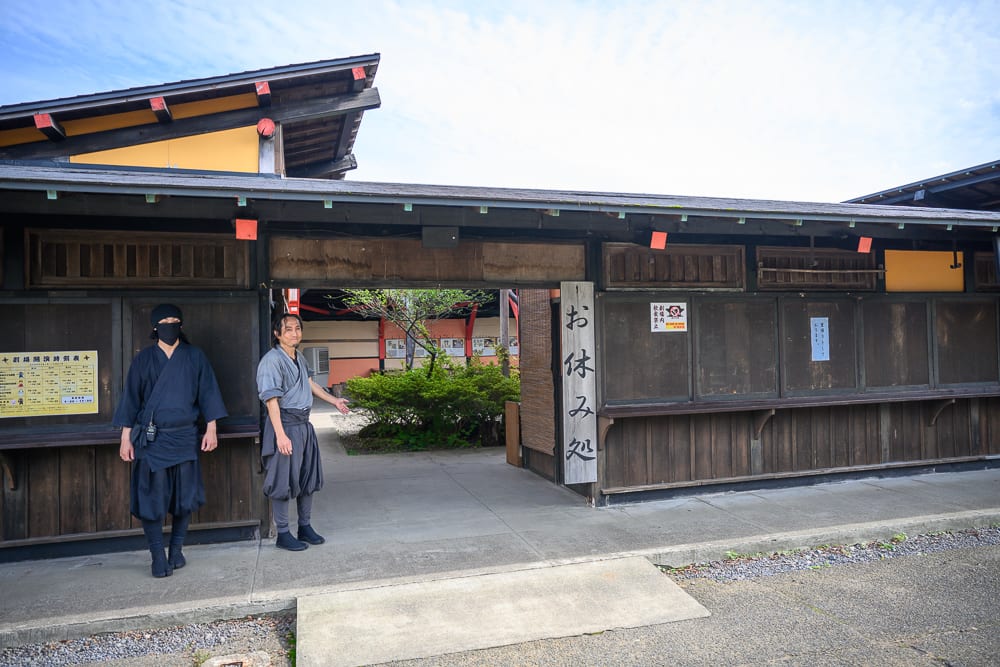 日本登別伊達時代村完整指南|江戶街景與庭園展示 9 日本登別-依達時代村|日式木造建築、木牆、黑衣男子、招牌、中庭、樹木,兩名身著傳統服飾的男子站在木造房舍外,門口懸掛直式日文招牌,中央通道可見綠色植物與建築結構。