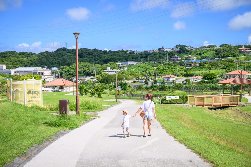 沖繩中城中城公園｜綠地、步道、木橋、路燈、房屋、山丘、藍天。成人牽著小孩走在戶外柏油路面上，兩旁為大片草皮與矮灌木叢，遠方有零星建物和電線橫跨天際，上方佈滿藍天白雲。