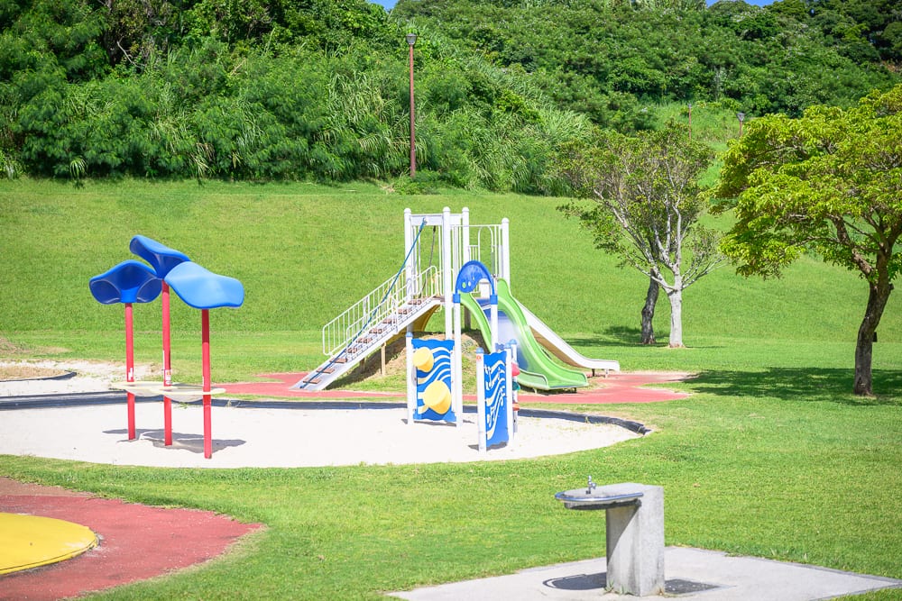 沖繩中城中城公園｜草地、滑梯、沙坑、樹木、路燈、飲水台，戶外公園內設置彩色遊樂設施，背景為綠色山坡，前方有水泥製飲水台，地面鋪設沙子與草皮。