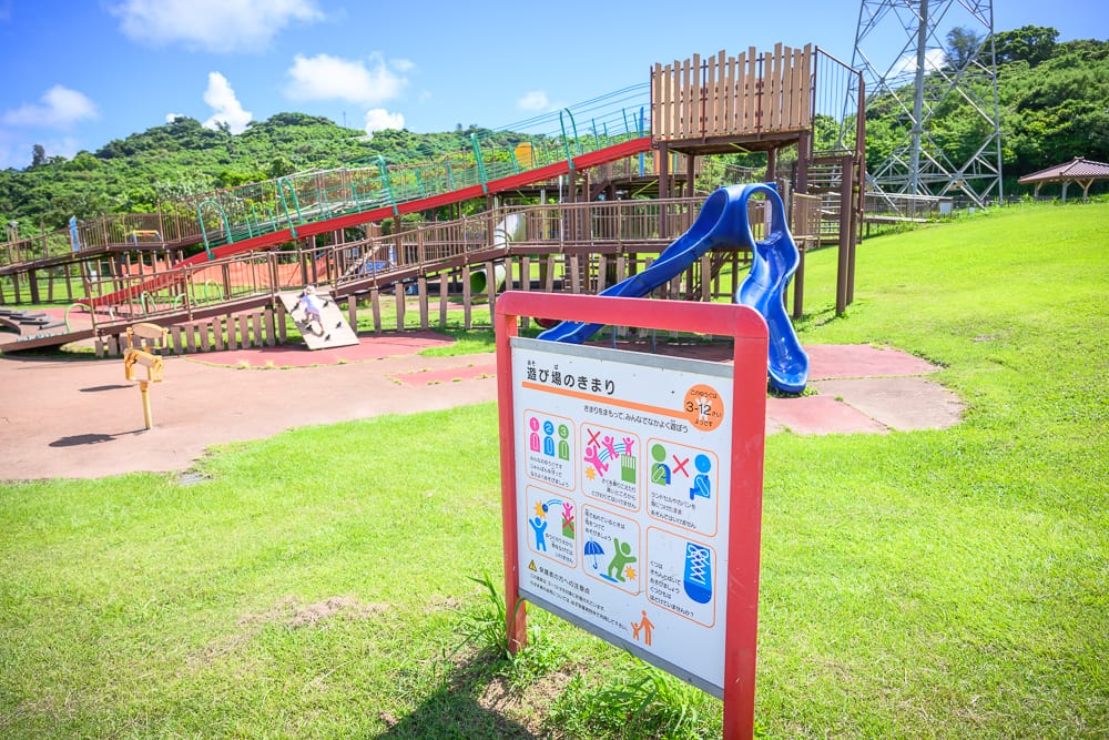 沖繩中城中城公園｜草地、指標牌、溜滑梯、遊具設施、山丘。藍天白雲下，戶外公園設有多層木造遊樂設施與藍色、紅色溜滑梯，前景立著紅色日文告示牌，後方為綠意山坡與鐵塔。