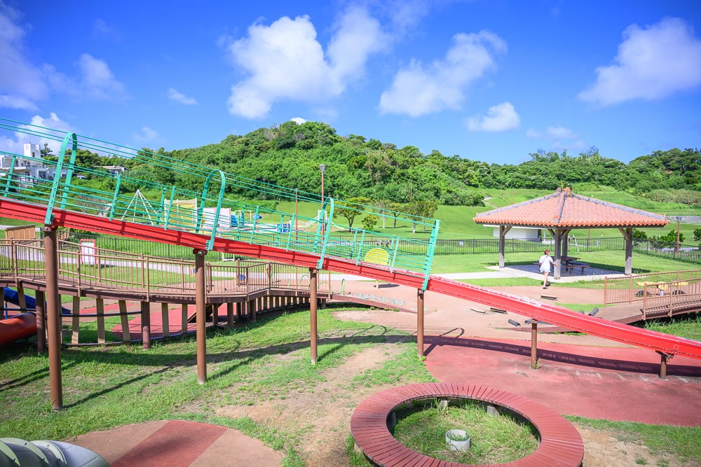 沖繩中城中城公園｜戶外公園、紅色長滑梯、綠地、涼亭、山丘、藍天白雲。紅色長滑梯貫穿畫面，遠處有綠意山丘與傳統涼亭，藍天白雲下草皮鋪展，視野遼闊。