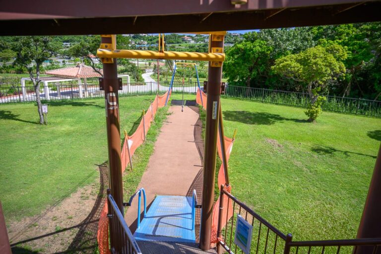 沖繩中城中城公園｜高處視角，溜滑索、跑道、草地、樹木、涼亭。褐色金屬柱支撐設施架構，橘色防護網圍繞軌道兩側，藍色金屬階梯與扶手位於前方，綠草地向遠方延伸，遠處可見白色建築與綠樹。