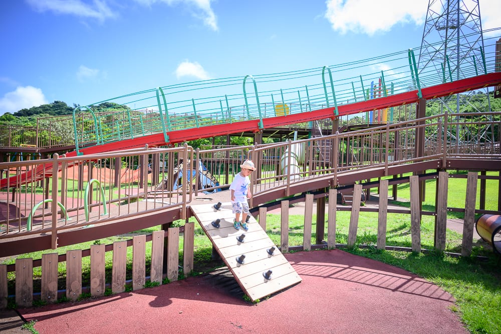 沖繩中城中城公園｜戶外公園、遊樂場、兒童、木棧道、溜滑梯、攀爬板、綠地、藍天。一名戴帽幼童站在木質斜坡上，周圍環繞紅色斜坡護欄、金屬高塔與綠樹，地面鋪設紅色彈性地墊。