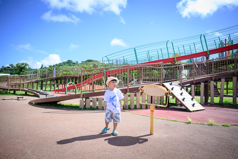 沖繩中城中城公園｜兒童、遊樂場、溜滑梯、天空、藍天白雲、樹木。戴著帽子的孩童站在紅色與木製的大型遊樂設施前，地面為紅棕色鋪面，身後有延伸的欄杆與樓梯。