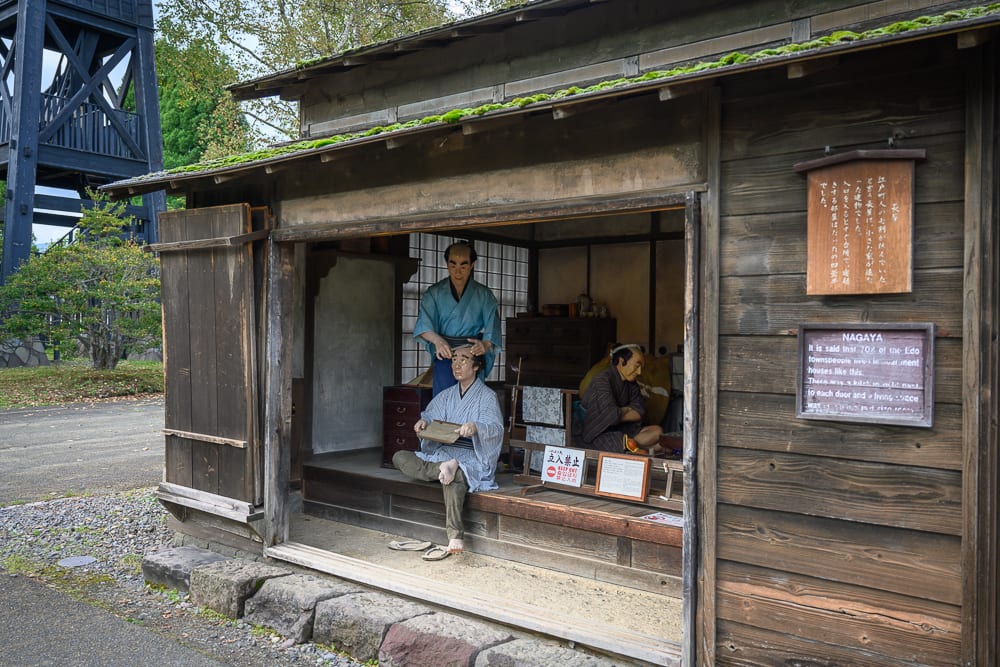 日本登別伊達時代村完整指南|江戶街景與庭園展示 5 日本登別-依達時代村|木造房屋,室內,人體模型,江戶時代,告示牌,草履,木造建築,戶外。傳統木造平房展示歷史場景,內部摆設蠟像模擬生活與理髮情景,牆面懸掛解說木牌。