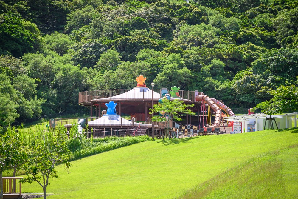 沖繩中城中城公園｜綠草地、溜滑梯、遊具設施、樹林、山坡，戶外兒童遊樂場座落於茂密綠樹環繞的山腳下，斜坡草皮向下延伸連接遊具平台，彩色塑膠溜滑梯與圓形遮陽棚構造清晰可見。