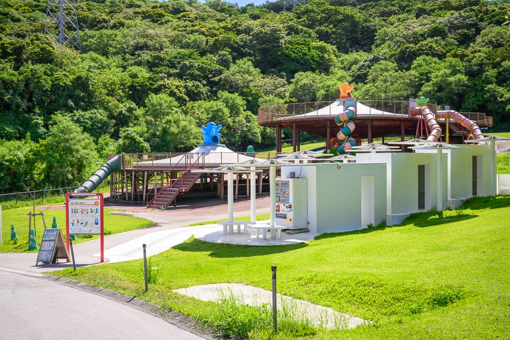 沖繩中城中城公園｜綠地、白色建物、自動販賣機、階梯、溜滑梯、遊樂場、圓形涼亭、樹林、鐵塔。綠地前方有一排白色立方體建築與自動販賣機，中段為木造平台與滑梯設施，後方及遠處為密集的山林綠樹。
