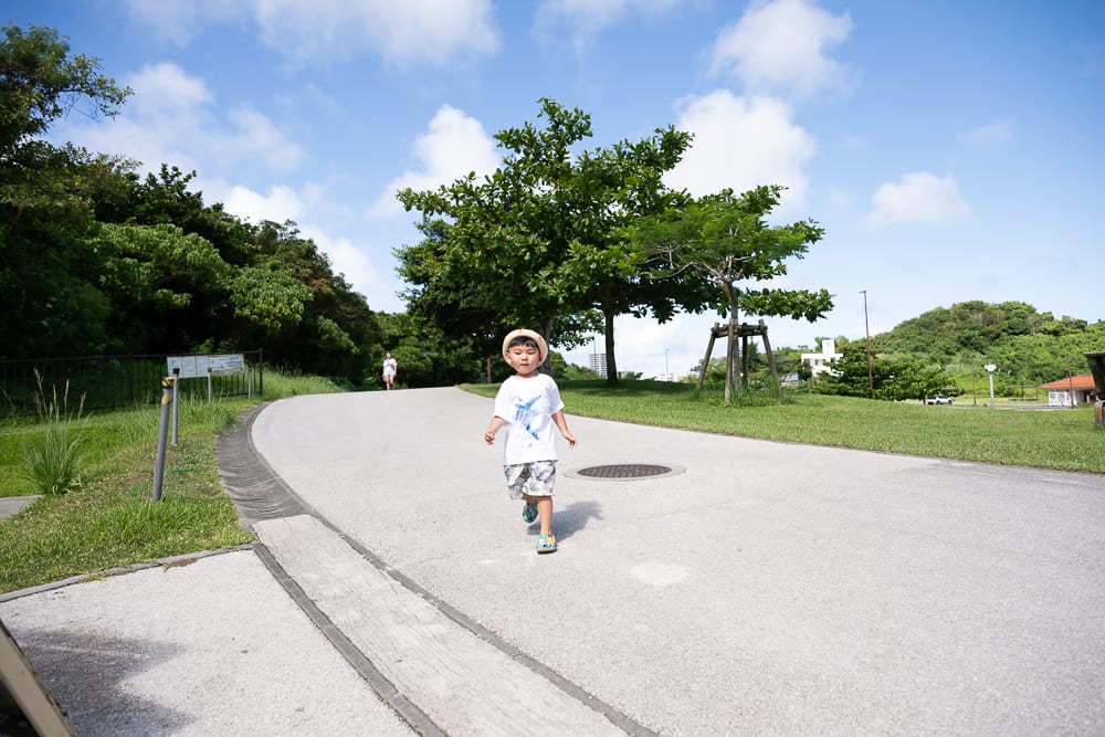沖繩中城中城公園｜柏油路、小孩、綠樹、草地、藍天，頭戴草帽與穿白衣的幼童在戶外鋪面道路上奔跑，兩側為草皮與樹木，背景為藍天與遠方山丘。