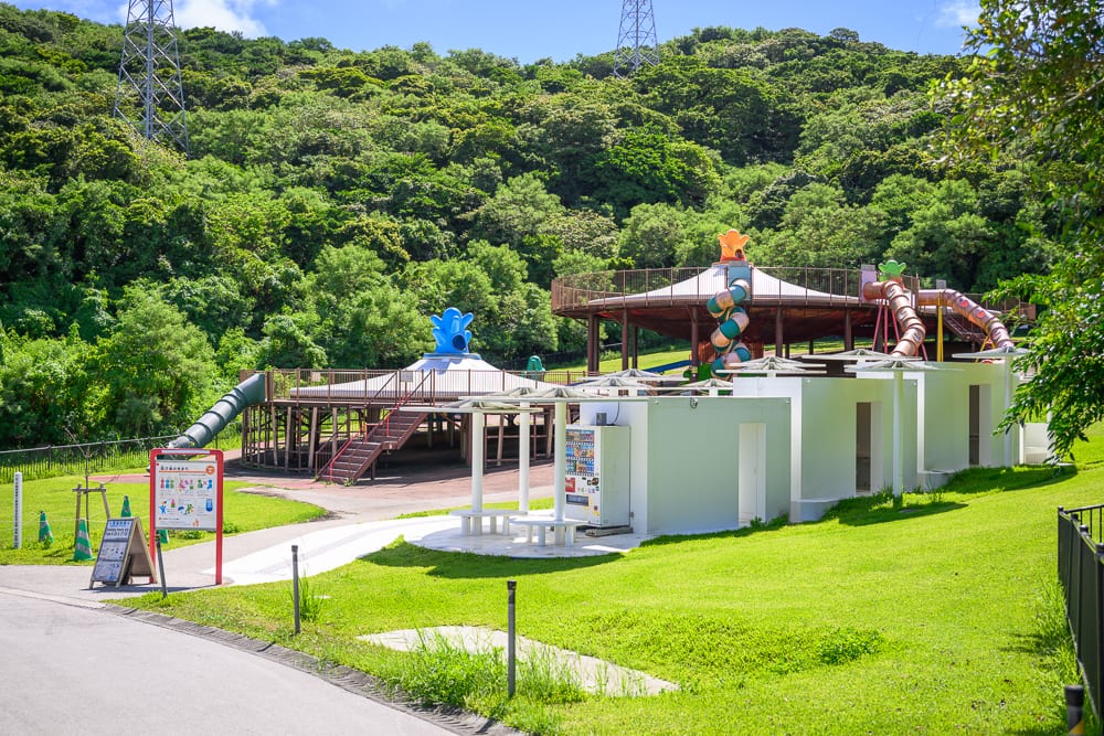沖繩中城中城公園｜綠草地、白色建築、溜滑梯、告示牌、自動販賣機、樹林、鐵塔、藍天。山腳下的戶外遊樂場，白色矮房旁設有休憩桌椅與販賣機，後方為木造設施與大型滑梯，遠方山坡密布綠樹並立有高壓電塔。