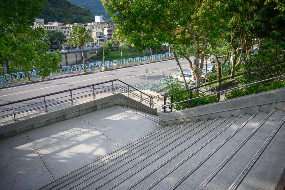 新北坪林茶業博物館完整指南|茶業歷史展覽與品茗體驗 13 新北坪林茶業博物館|戶外階梯、街道、橋、河、樹木、房屋,水泥階梯延伸至馬路,對面設有藍色欄杆的河道與低矮建築,遠方為綠意山巒。