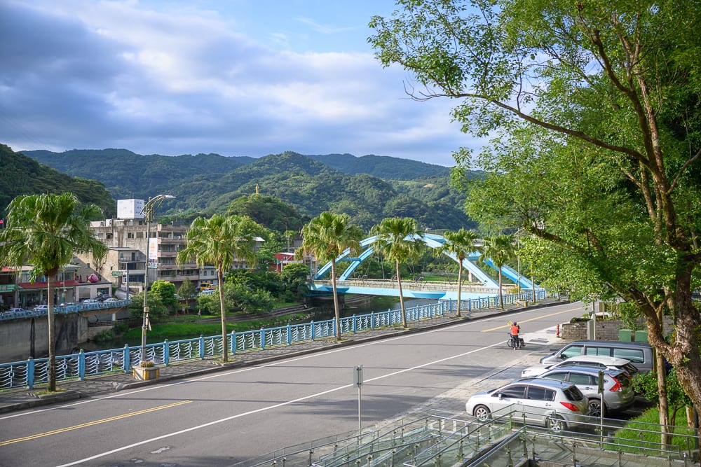 新北坪林茶業博物館完整指南|茶業歷史展覽與品茗體驗 6 新北坪林茶業博物館|橋、河、街道、椰子樹、汽車、山脈、建築物、雲朵,藍色拱橋橫跨河面,兩側道路停放數輛轎車,路旁整排椰子樹延伸至遠方,遠處為綿延綠山與多雲天空。